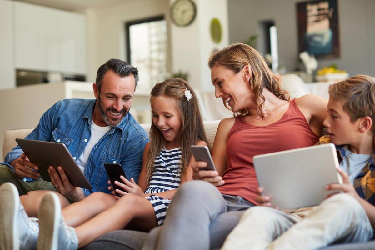 happy family of four at home smiling on the couch together with their devices