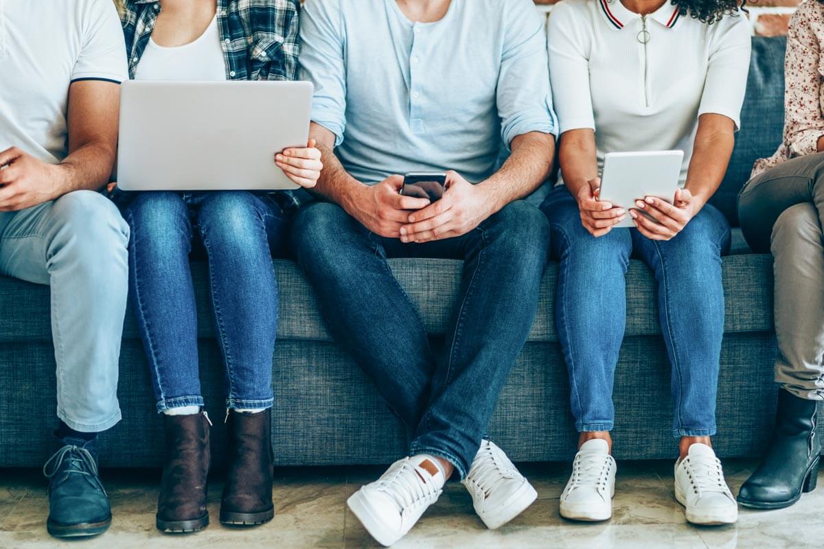 row of people sitting on couch holding various electronic devices