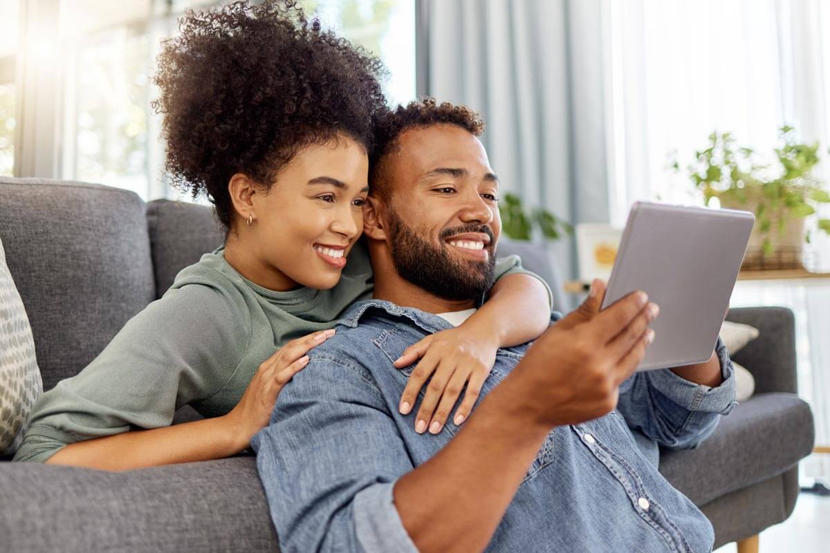 man and woman smiling and looking at tablet while laying on couch together